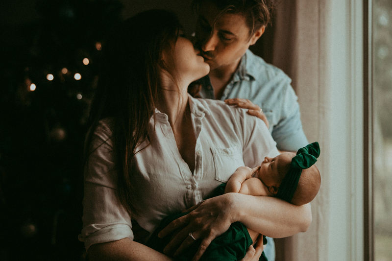 A couple kissing while the wife holds their newborn baby in front of a softly lit christmas tree and the soft light of the window they are standing beside captured by a Frederick Maryland Newborn photographer