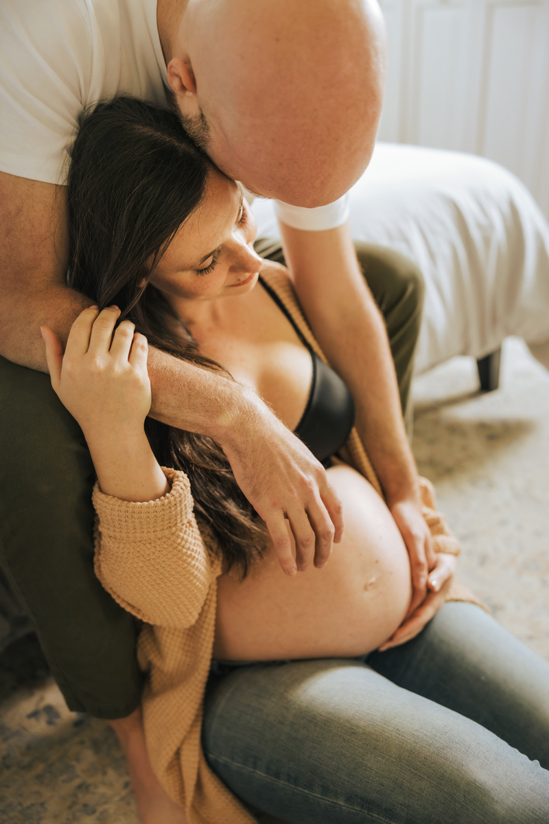 A man sitting on the edge of a bed leaning over to embrace his pregnant wife and her belly while she sits on the floor captured by a Cumberland Maryland maternity photographer