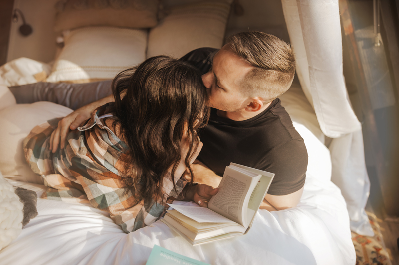 A man tenderly kissing the top of his wife's head as they snuggle in bed reading together captured by a Winchester Virginia elopement photographer